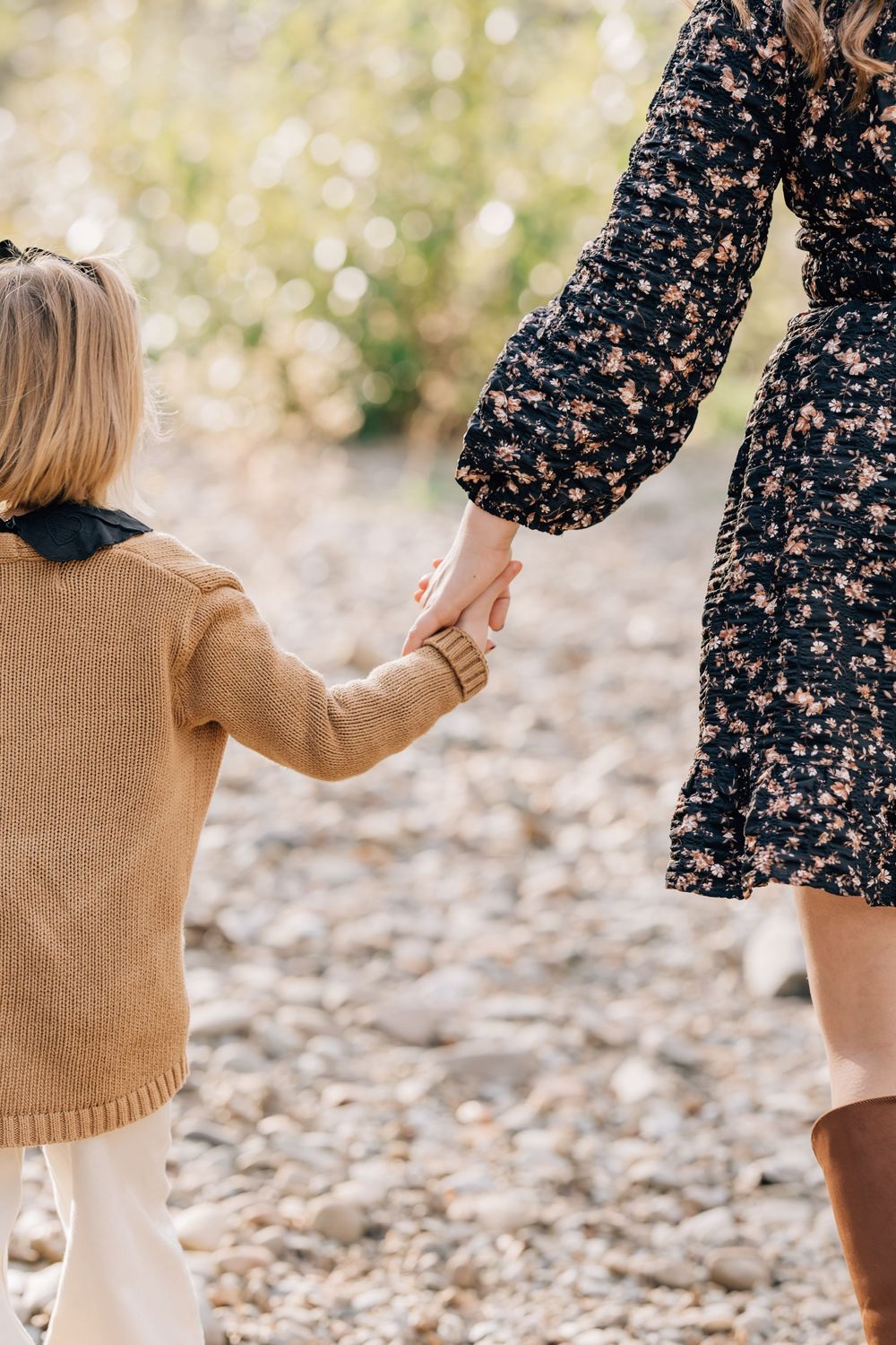 A woman in a floral dress holds hands with a young child in a tan sweater while walking outdoors.