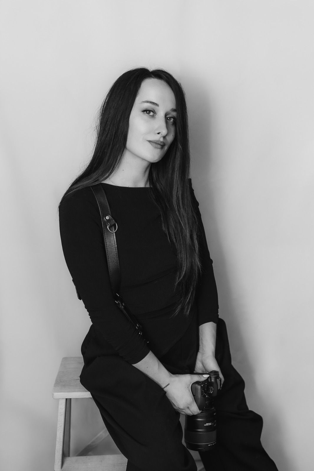 Young woman with long red hair in black outfit holding a DSLR camera, seated on wooden stool against gray background.