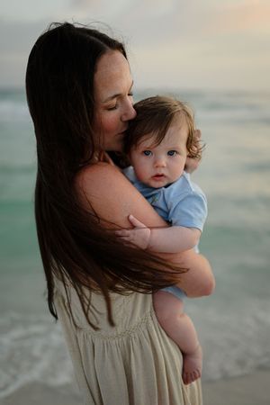 A mother tenderly kisses her baby on a beach at sunset, waves and turquoise water in the background.