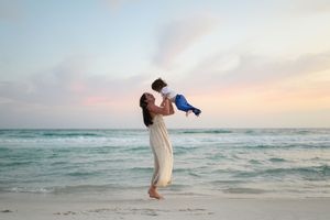 Mother lifting baby joyfully on a serene beach at sunset, wearing a white dress with ocean waves behind.