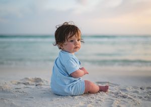 A cute baby in a blue romper sits on a sandy beach with turquoise ocean waves in the background.