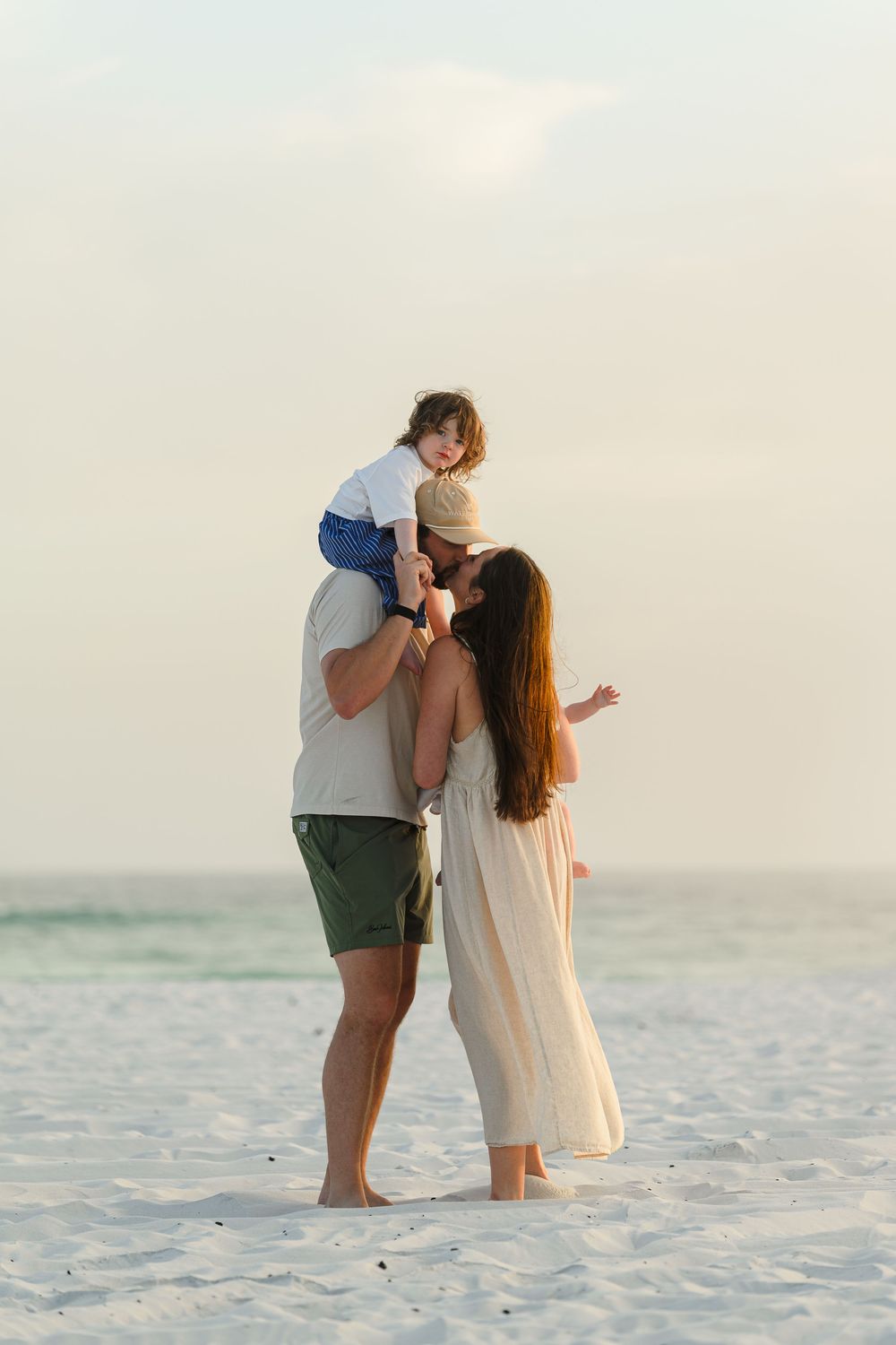Family of three enjoying a sunset beach moment, with dad holding toddler on shoulders while mom embraces them on white sand.
