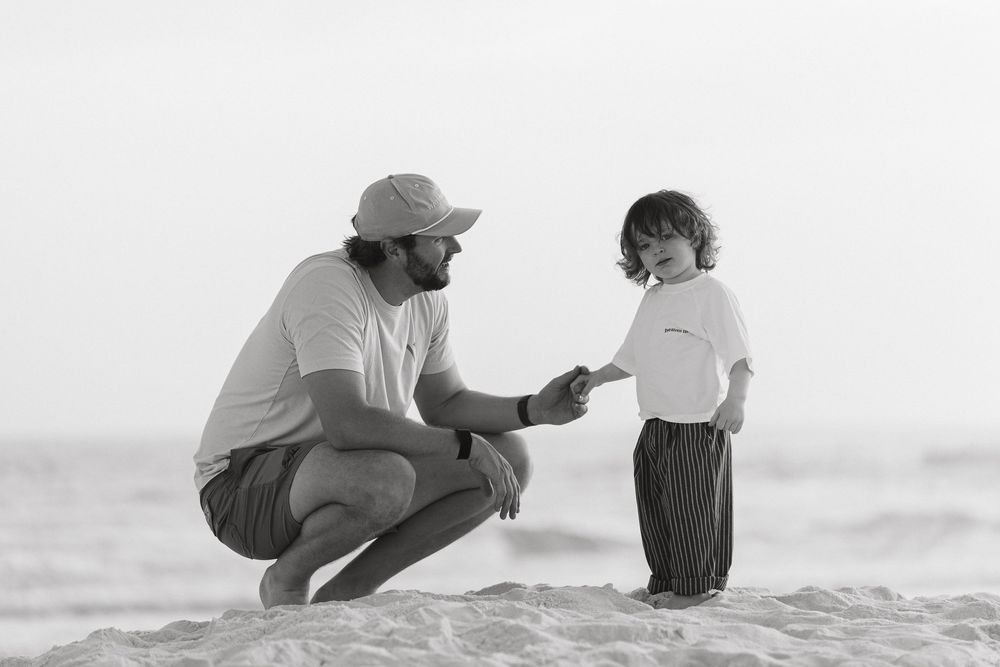Black and white photo of a father crouching on a sandy beach, sharing a tender moment with his young toddler child.