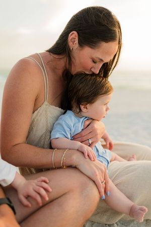 A mother tenderly kisses her baby boy on the head while sitting on a sandy beach at sunset.