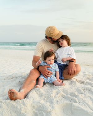 A father sits on white sand beach holding two young children, with ocean waves in the background.