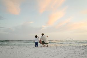 Adult crouching beside young child on sandy beach at sunset, pointing toward ocean horizon together.
