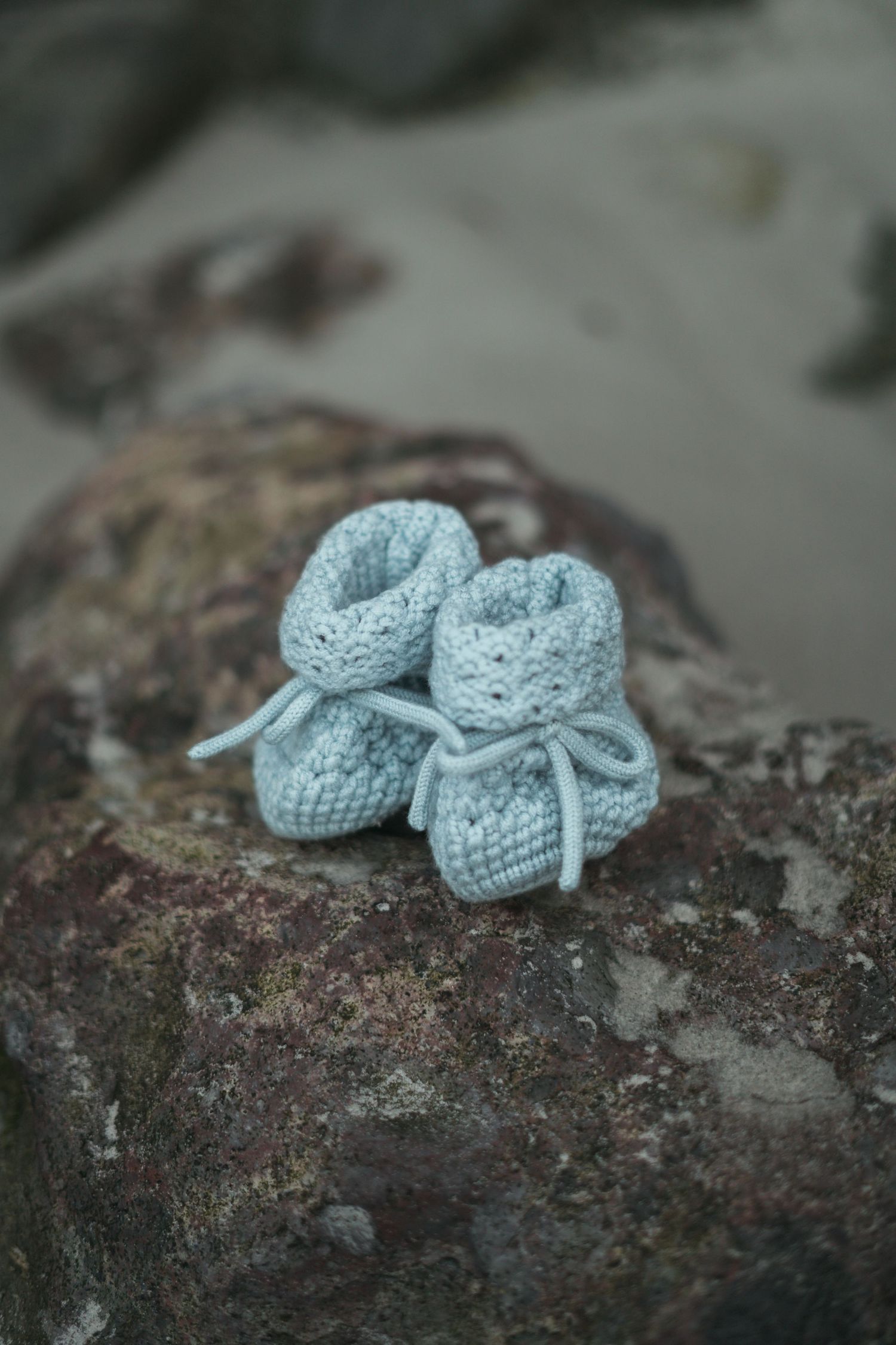 Tiny light blue knitted baby booties with bow ties resting on a mossy rock outdoors.