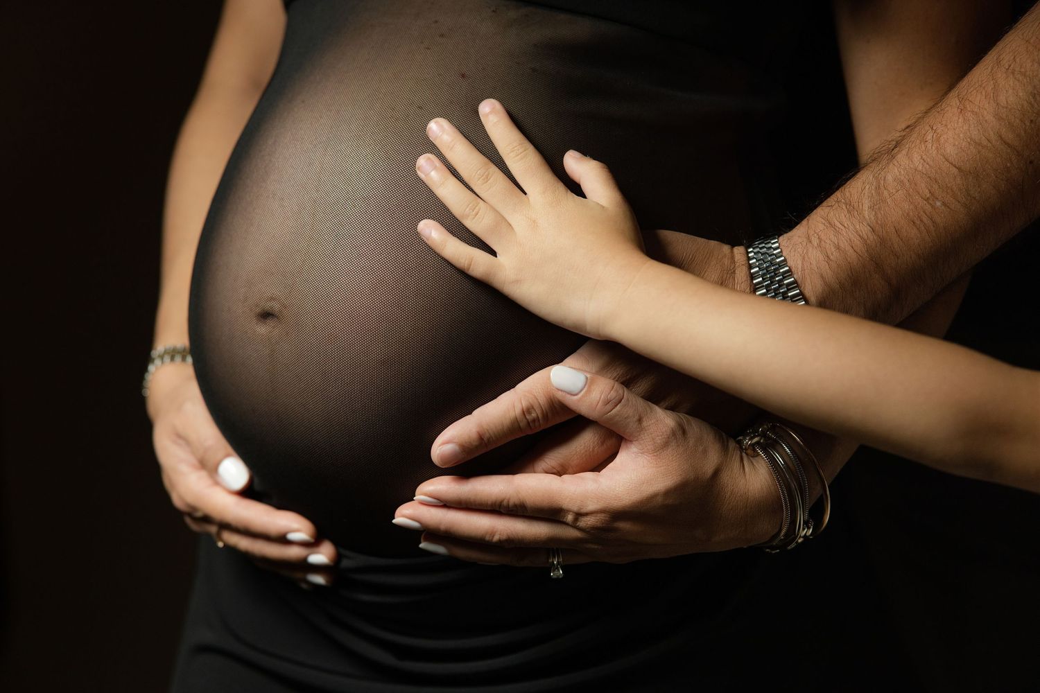 Pregnant woman in black dress with partner and child's hands gently resting on baby bump.