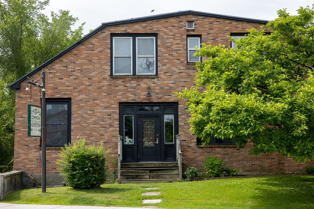 Two-story brick building with black front door, decorative glass, surrounded by mature trees and green landscaping.