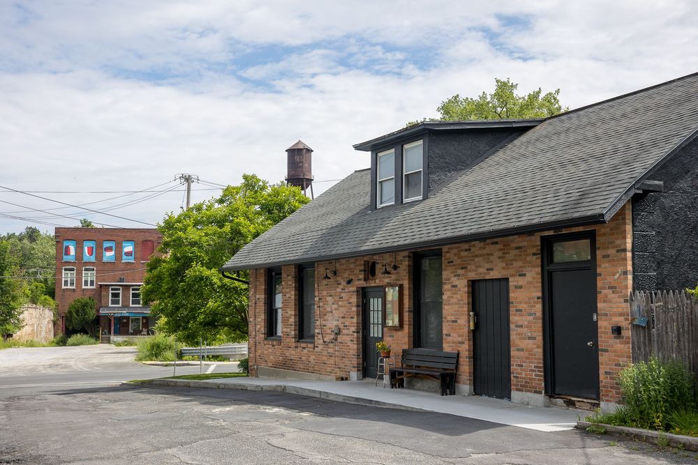Exterior of a brick building with dark roof, bench by entrance, parking lot, trees, and water tower in background.