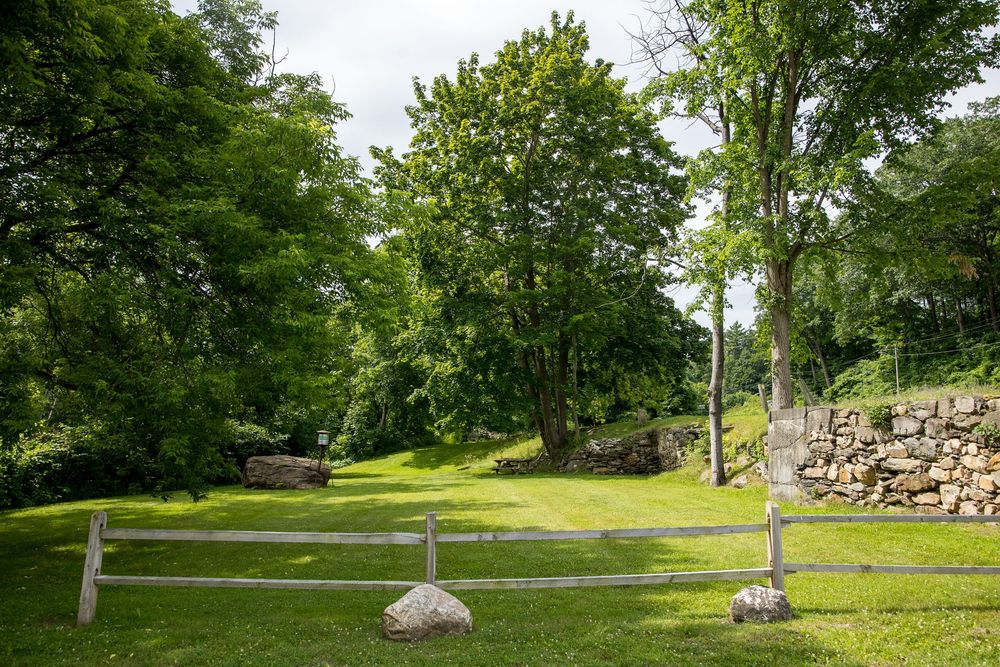 Serene green landscape with wooden split-rail fence, large trees, and rustic stone wall in background.