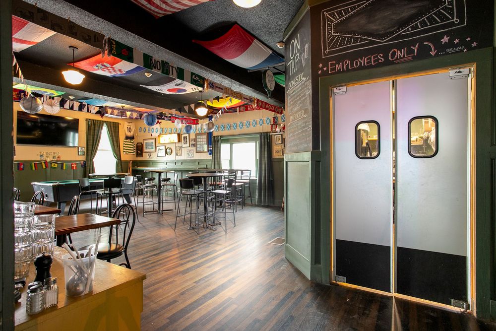 Sports bar interior with wooden tables, international flags, chalkboard signage, and employees-only double doors.