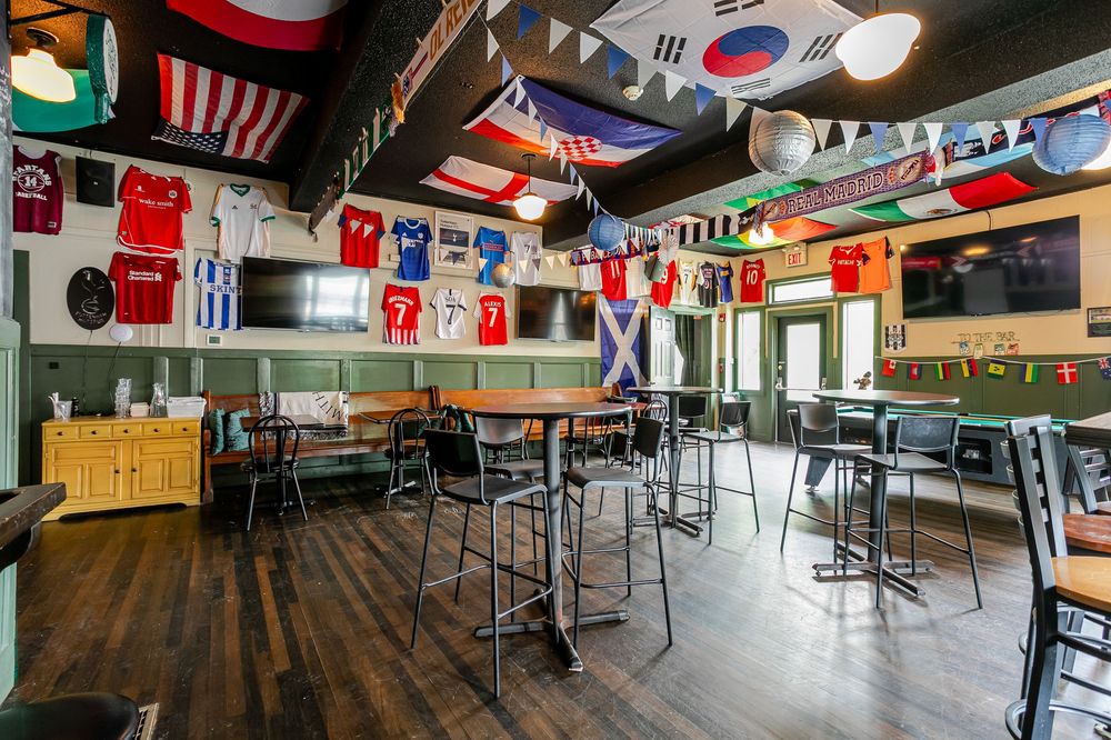 Sports bar interior decorated with international soccer jerseys and flags, featuring high-top tables and bar stools.