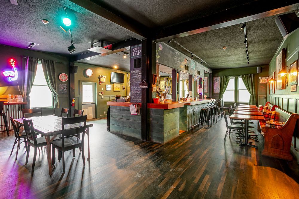 Spacious bar interior with dark hardwood floors, green walls, central bar, neon signs, and booth seating.