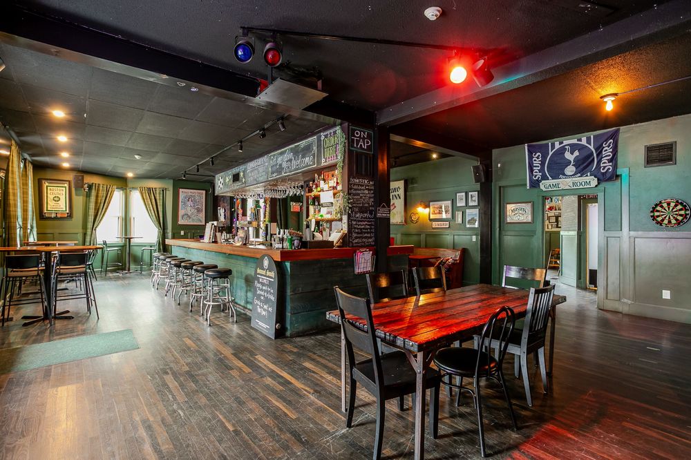 Dimly lit pub interior with wooden tables, bar with stools, dartboard, and moody green walls and dark ceiling.