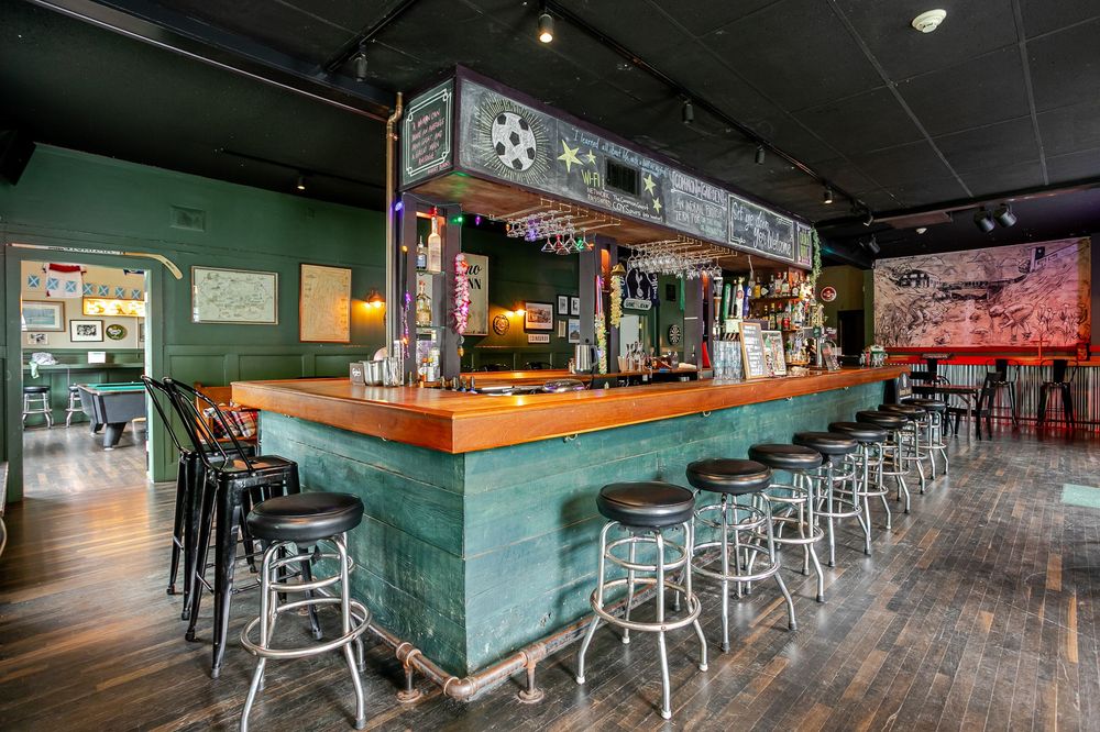 Teal-painted bar counter with wooden top, industrial pipe shelving, backlit bottles, and black bar stools.