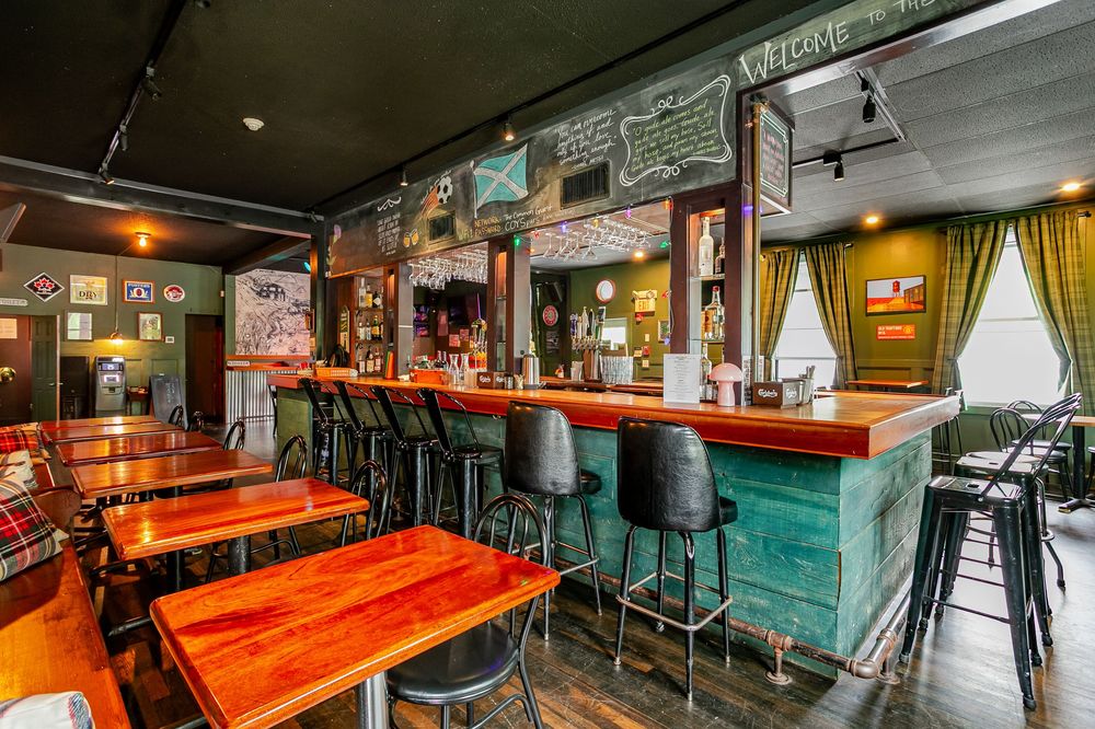 Cozy pub interior with wooden tables, teal bar, chalkboard menus, green walls, and warm ambient lighting.