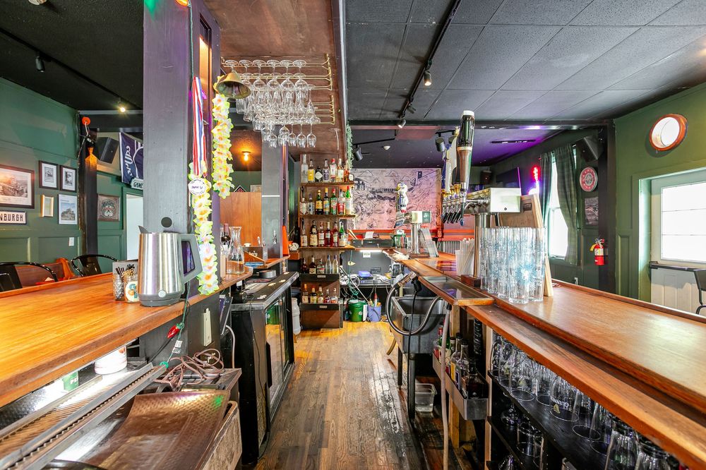 Behind-the-bar view showing wooden countertop, hanging wine glasses, bottles, and colorful bar decorations.