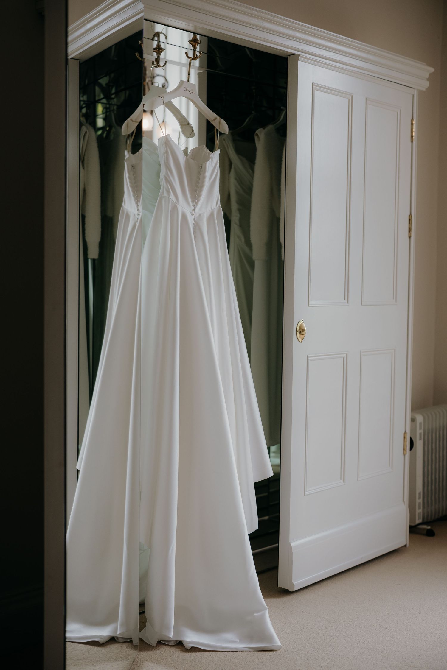 Elegant white satin A-line wedding dress hanging on a wardrobe door in a bright, classic bridal suite.