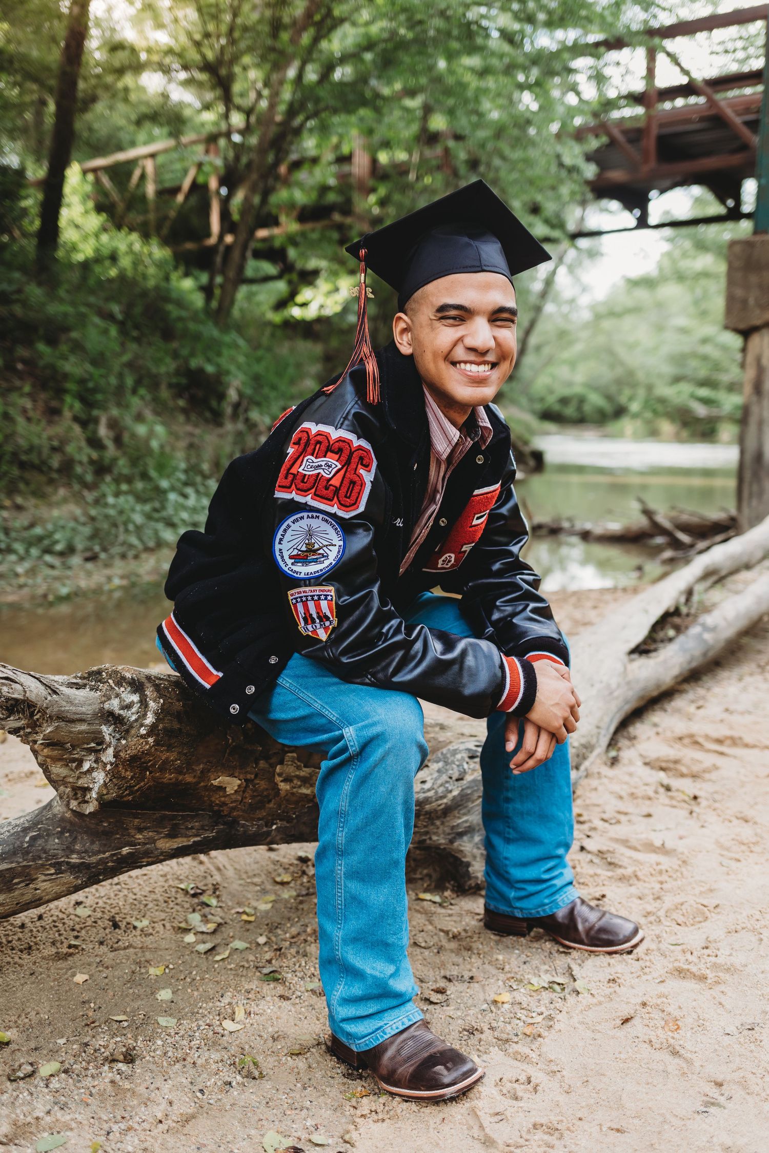 Smiling graduate in cap and letterman jacket sitting on a log outdoors near a creek.