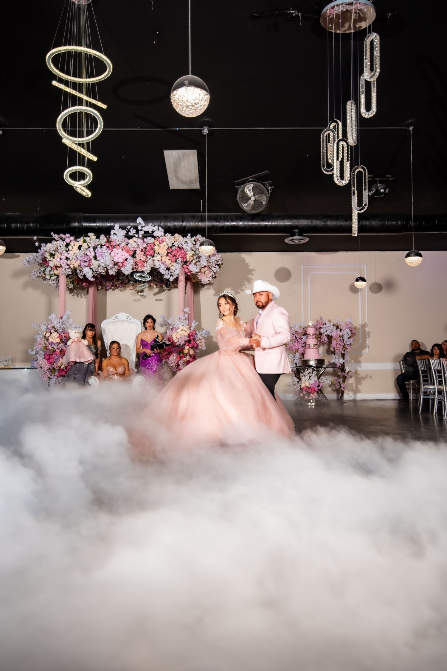 Quinceañera couple dancing in pink ballgown surrounded by dramatic fog on elegant venue dance floor.