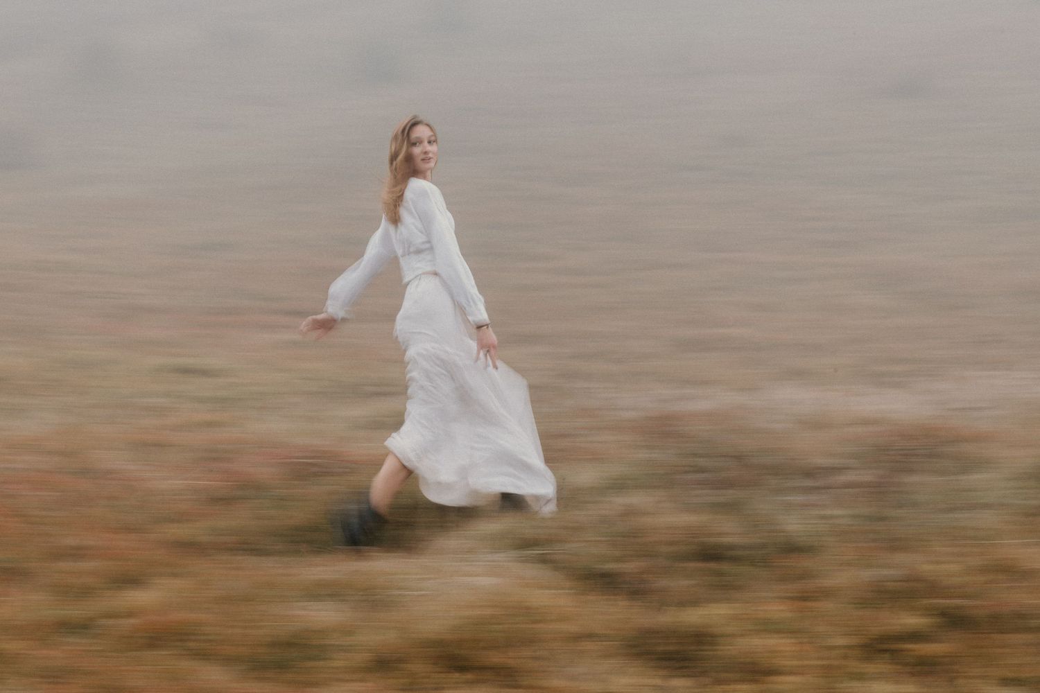 Woman in white dress standing on misty mountain hillside with fog rolling over golden brown landscape.