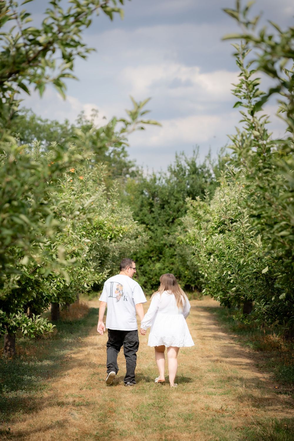 Couple holding hands walking through lush green orchard rows, woman in white dress, man in graphic tee, viewed from behind.