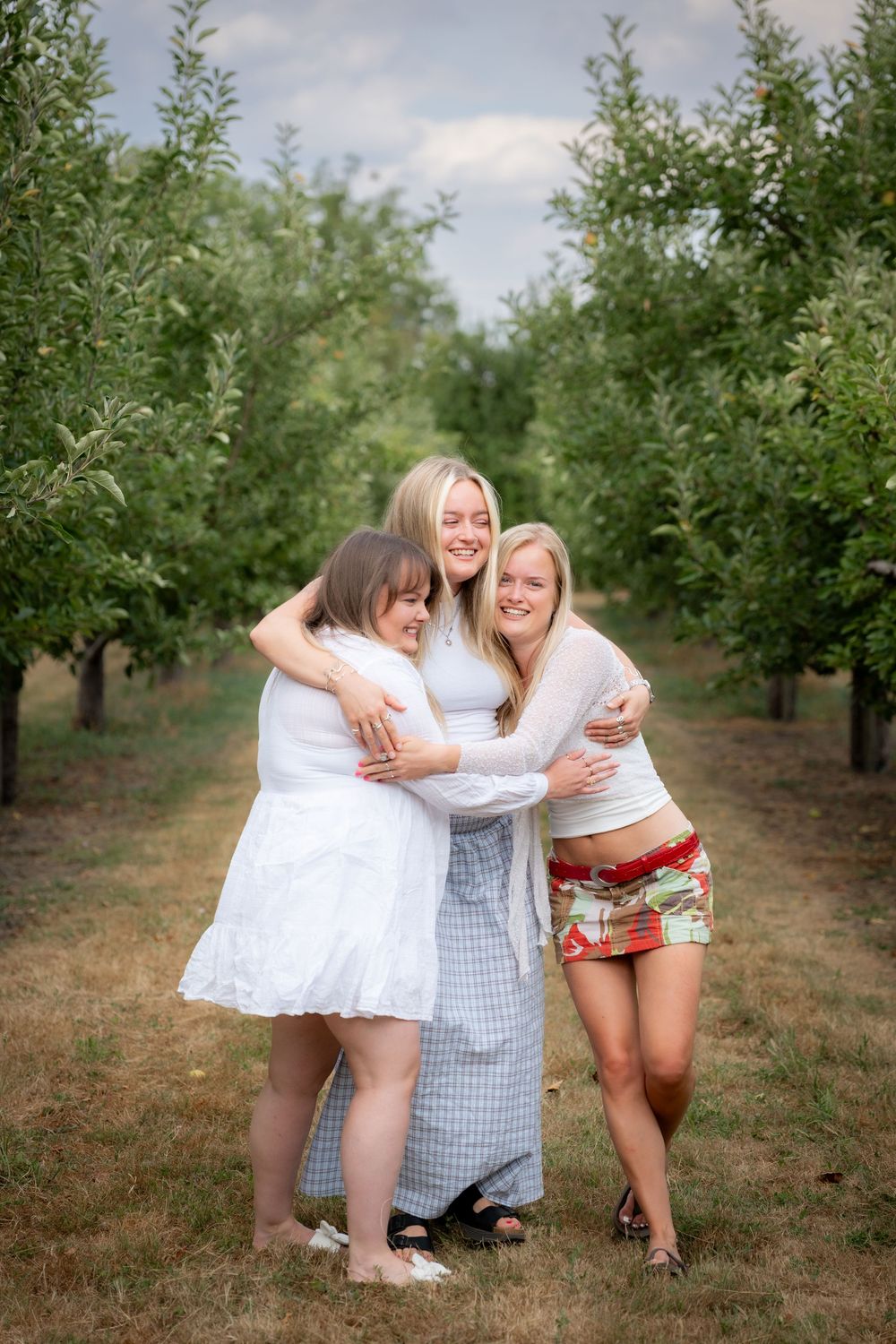 Three smiling young women hugging each other in a lush green orchard pathway on a cloudy day.