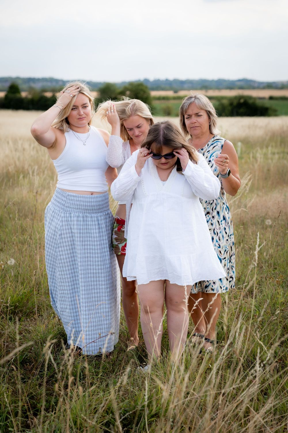 Four women in white and blue outfits pose together in a golden field, adjusting hair and sunglasses on a cloudy day.
