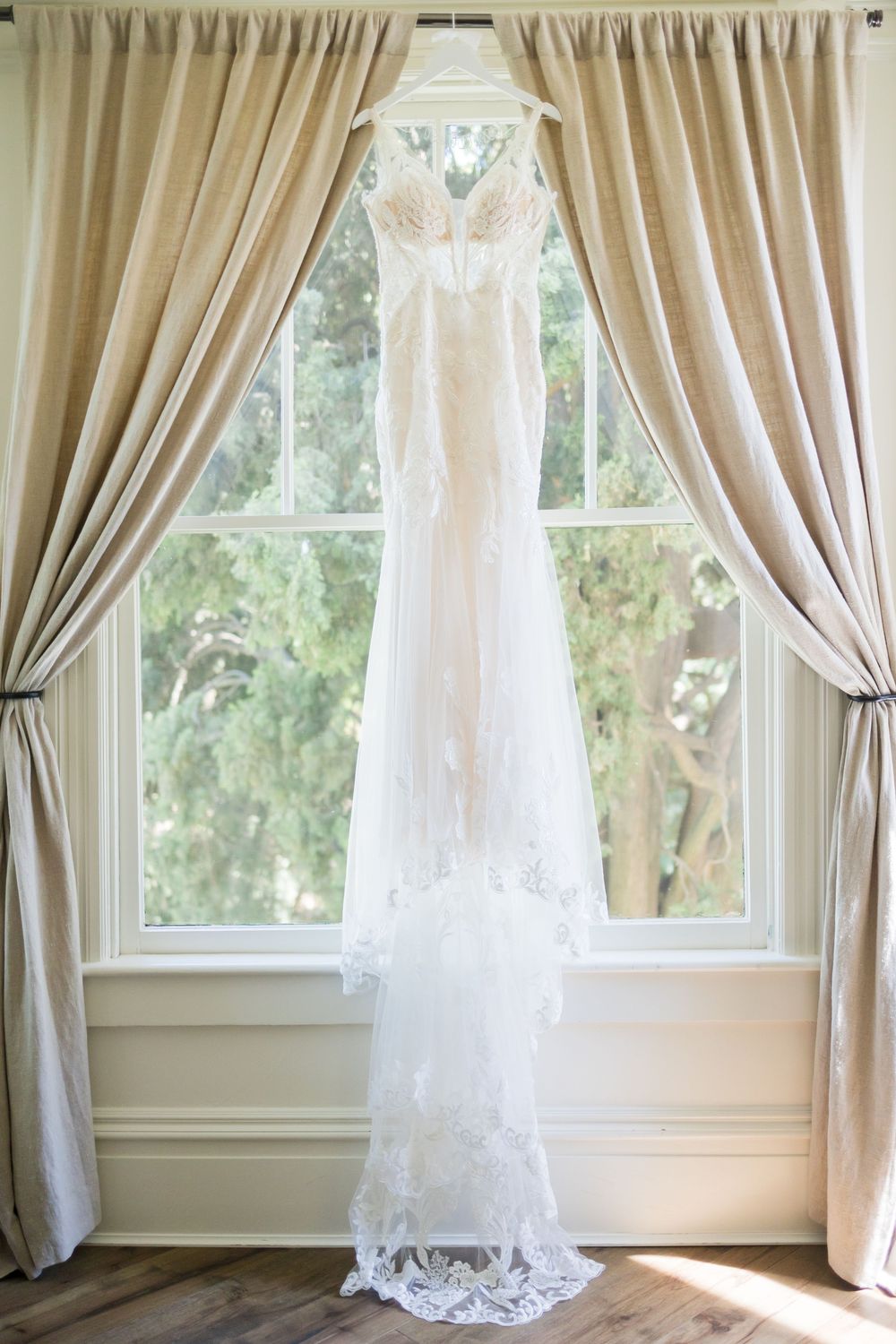 Elegant lace wedding dress hanging in a sunlit window with neutral drapes.