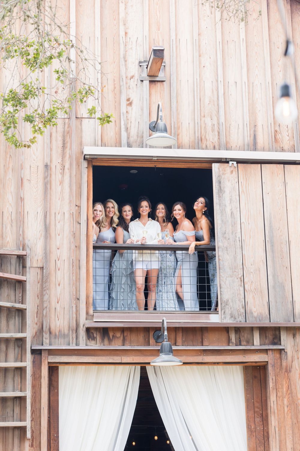 Bridesmaids in gray dresses with bride in white pose on rustic barn balcony at outdoor wedding venue.
