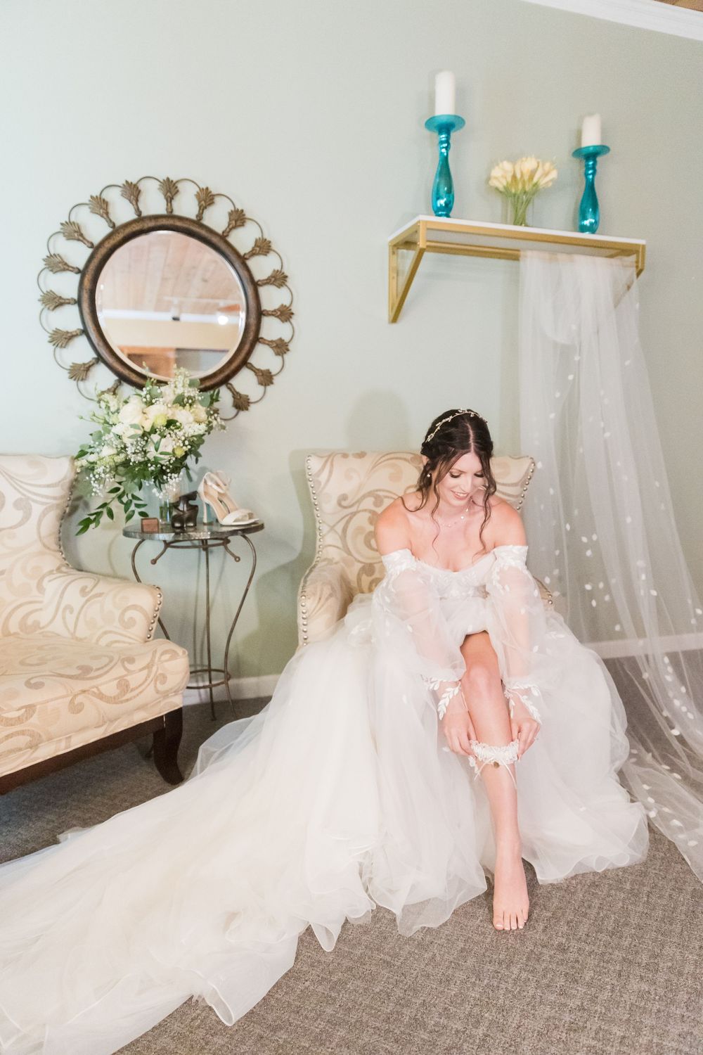 Bride in elegant white ball gown sitting in chair putting on garter in a softly decorated bridal suite.