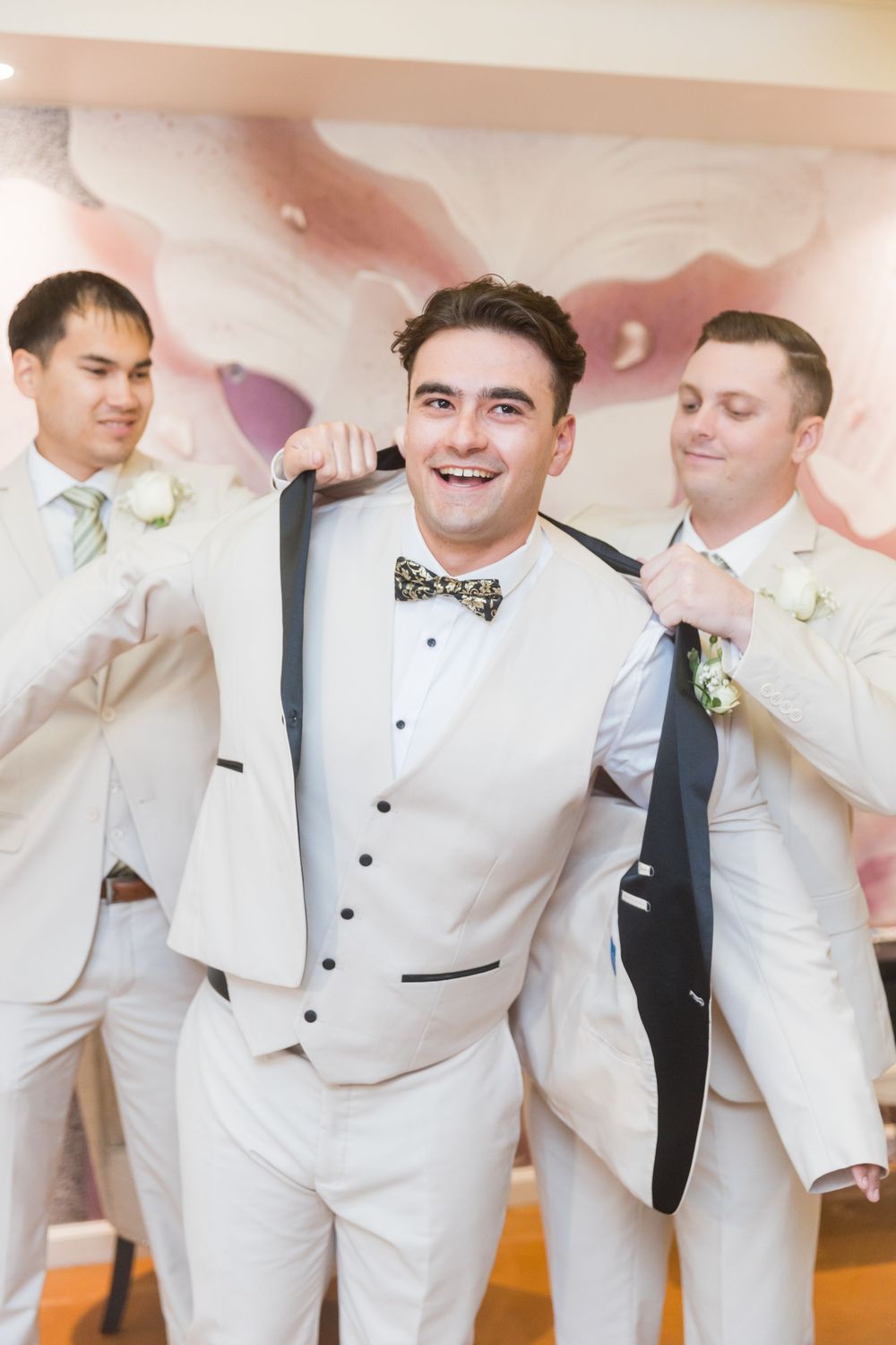 Groom smiling in white vest and bow tie as groomsmen help him into his suit jacket before the wedding ceremony.