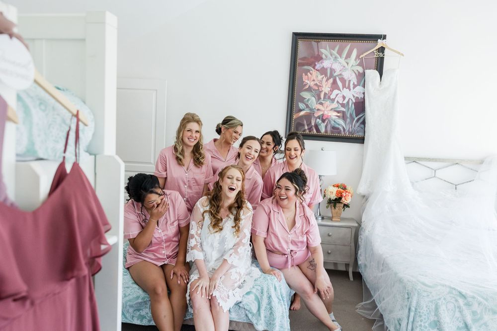 Bride and bridesmaids in pink robes laughing together in a bright bedroom before the wedding ceremony.