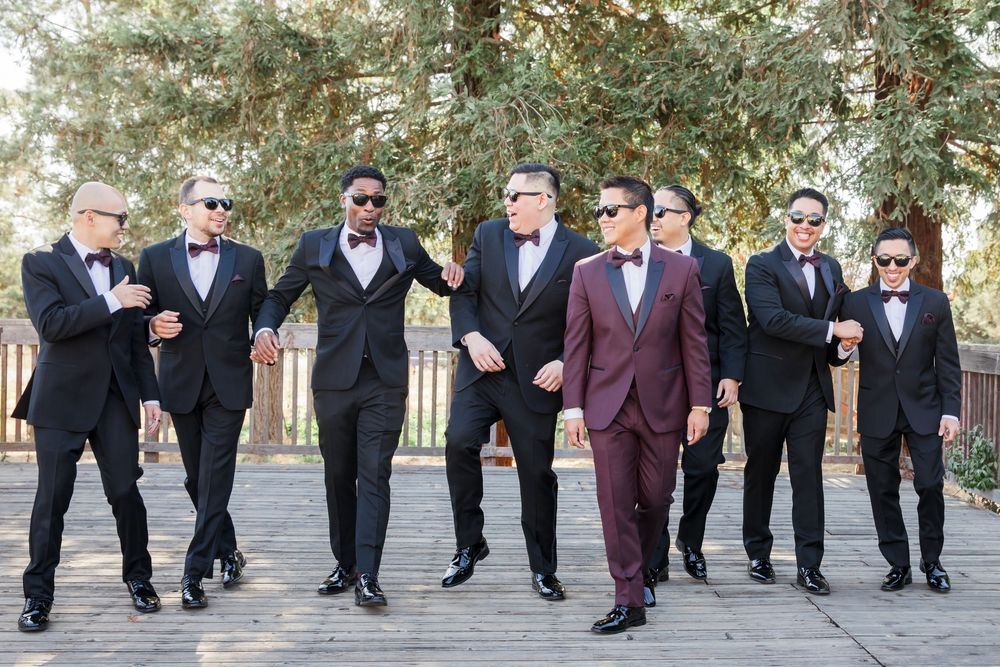 Groom in burgundy tuxedo poses with six groomsmen in black tuxedos and bow ties wearing sunglasses outdoors.
