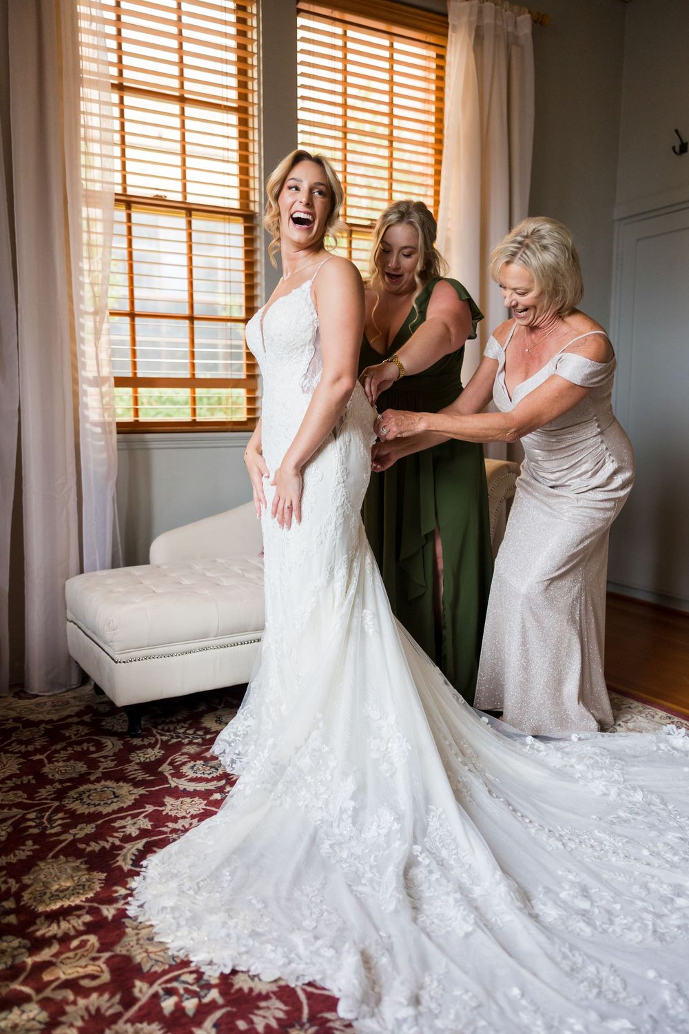 A bride laughs joyfully as a bridesmaid and mother help fasten her lace wedding gown in an elegant room.