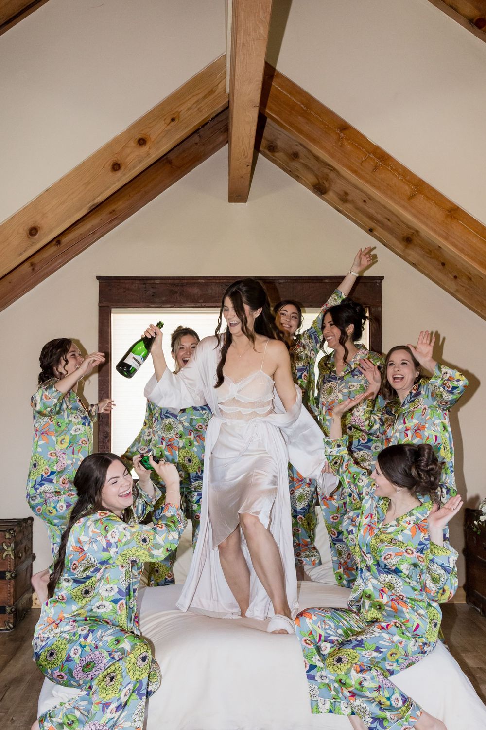 Bride in white robe jumps on bed surrounded by bridesmaids in floral pajamas celebrating with champagne.