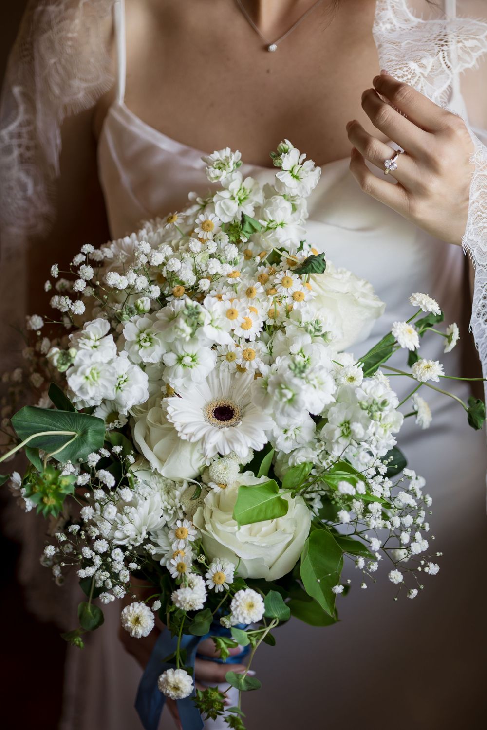 Bride holding a lush white wedding bouquet with gerberas, roses, daisies, and baby's breath, wearing a diamond engagement ring.