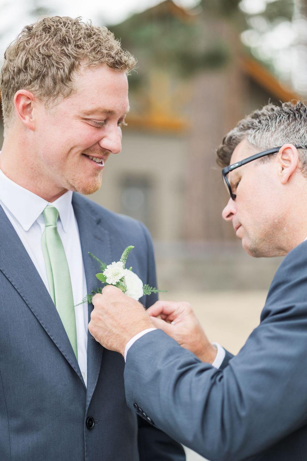 A man in a gray suit with green tie smiles as another man pins a white boutonniere on his lapel.