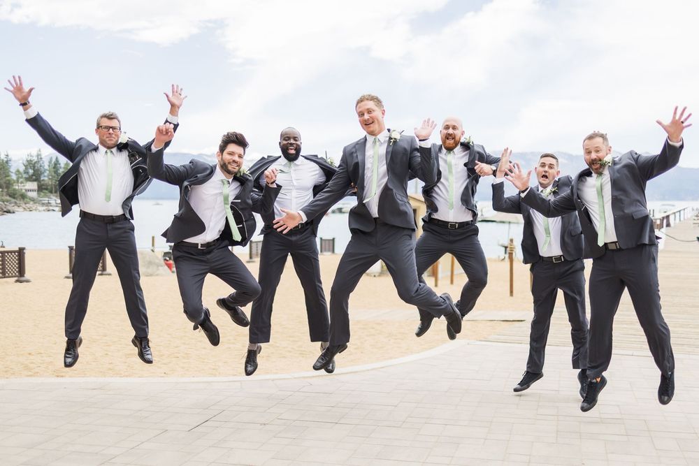 Groomsmen in black tuxedos joyfully jumping in unison outdoors at a beach wedding celebration.