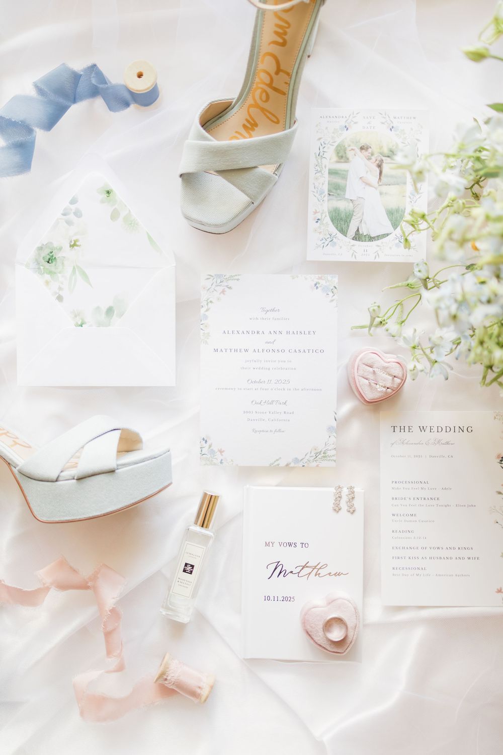 Elegant wedding flat lay with floral invitations, blue heels, pink ribbons, perfume, and delicate accessories on white fabric.