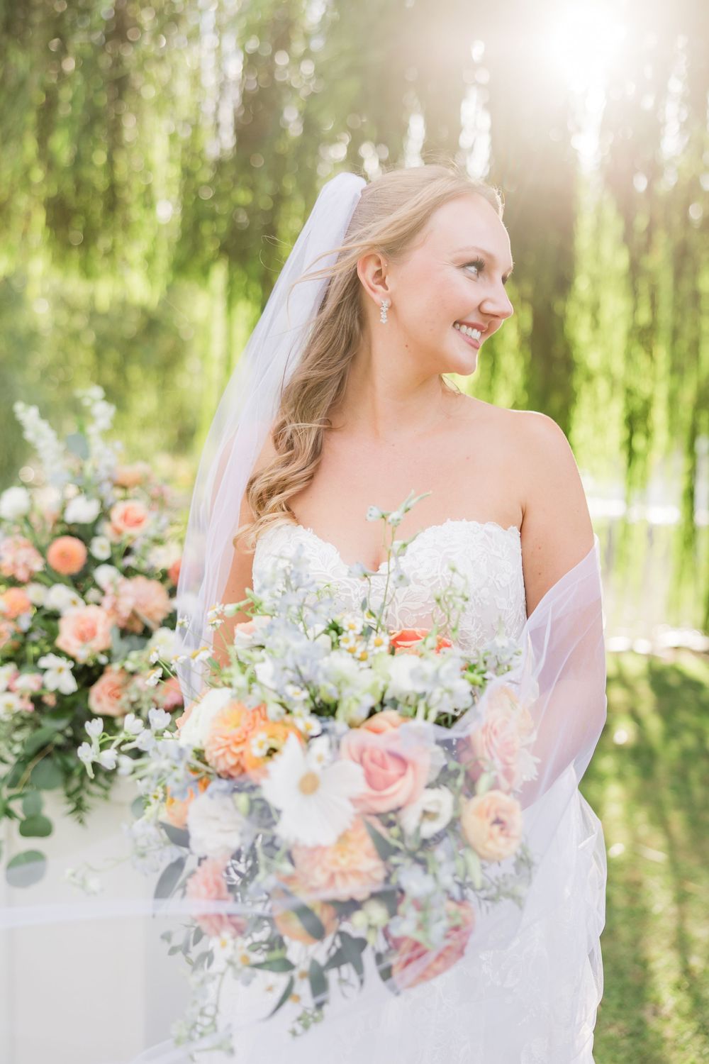 Smiling bride in strapless gown and veil holds a lush peach and white bouquet outdoors in soft sunlight.
