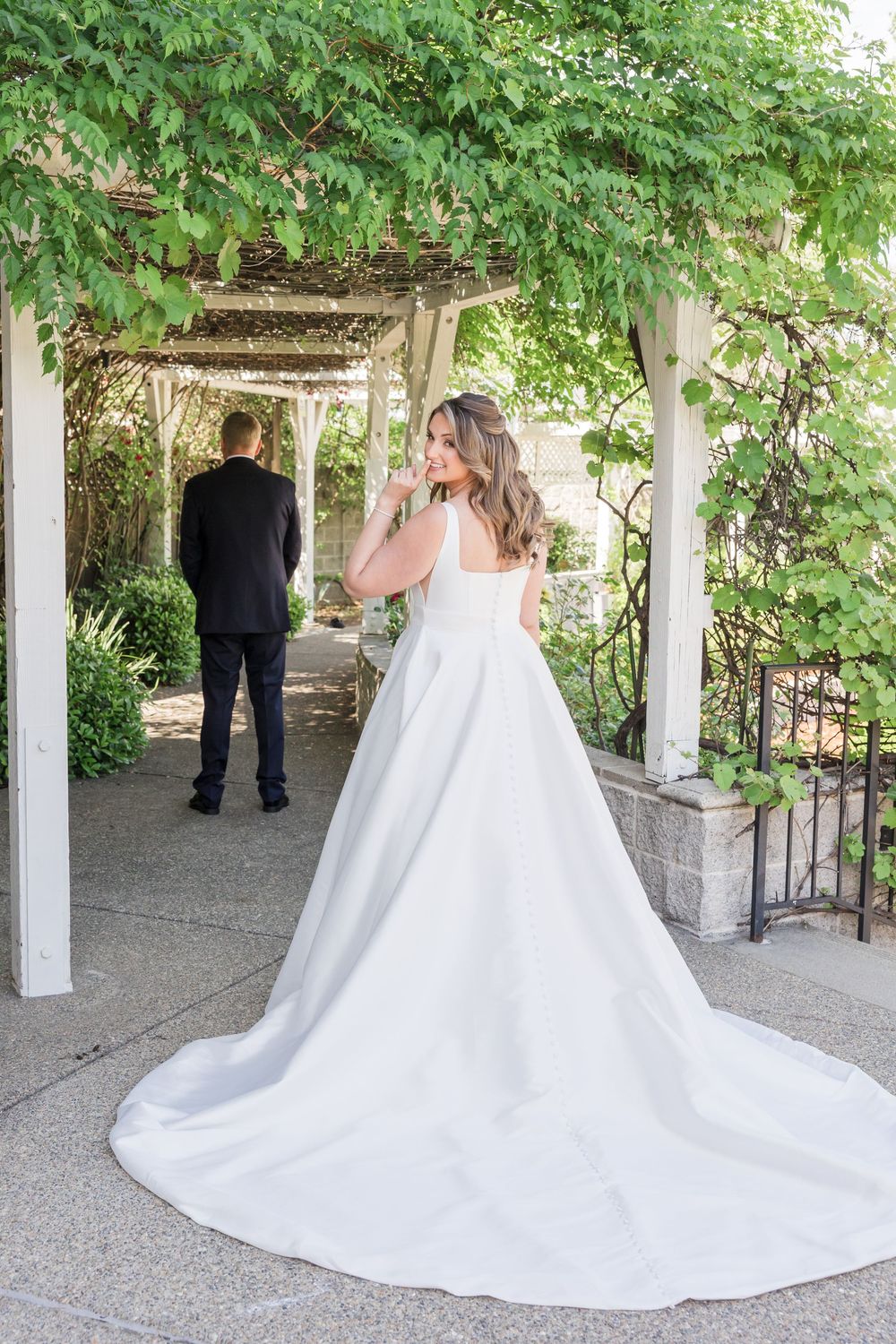 Bride in elegant white ball gown with long train stands under vine-covered pergola as groom in black suit walks ahead.