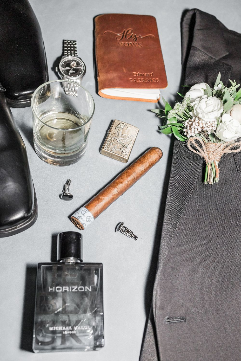 Groom flat lay featuring Horizon cologne, cigar, watch, cufflinks, boutonniere, leather journal, whiskey glass, and black dress shoes.