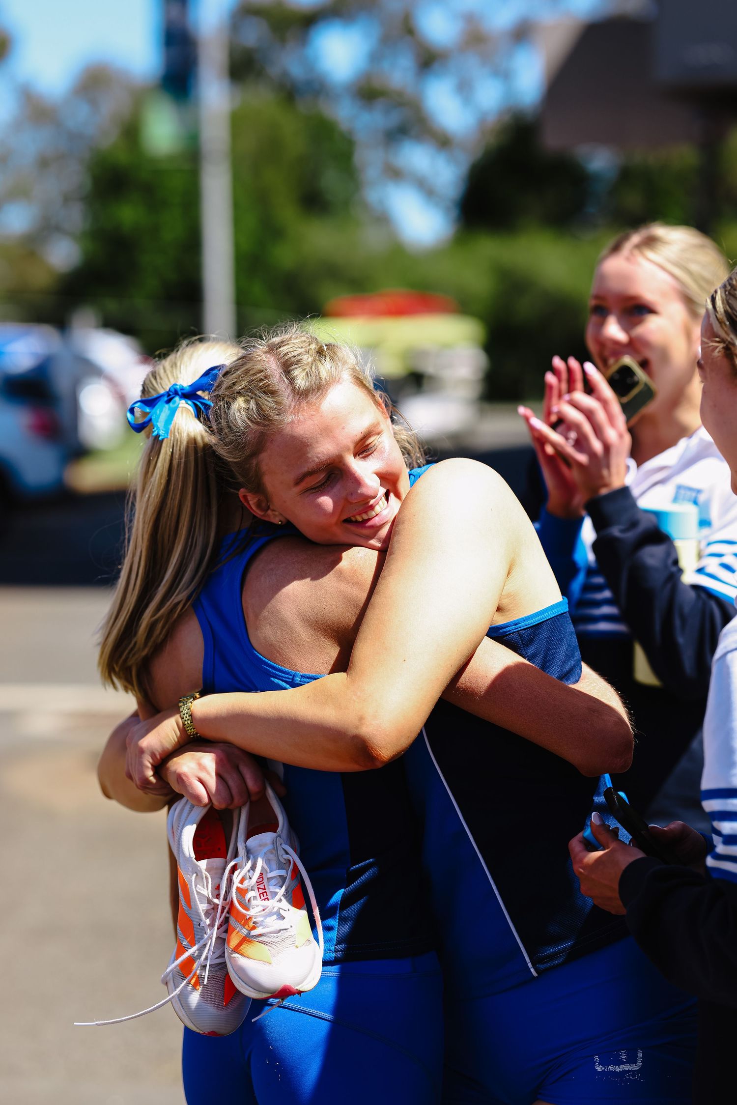 Two female athletes in blue uniforms share an emotional hug outdoors, while a teammate claps in celebration behind them.