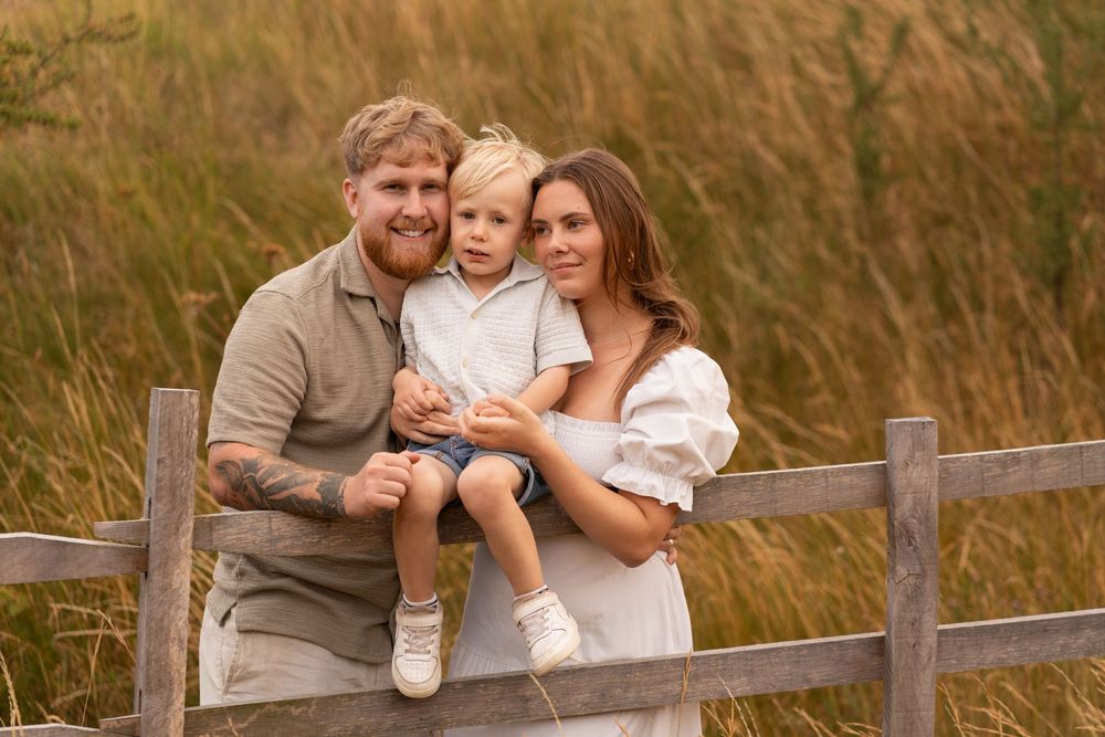 Happy family of three posing outdoors by a wooden fence with tall golden grass in the background.