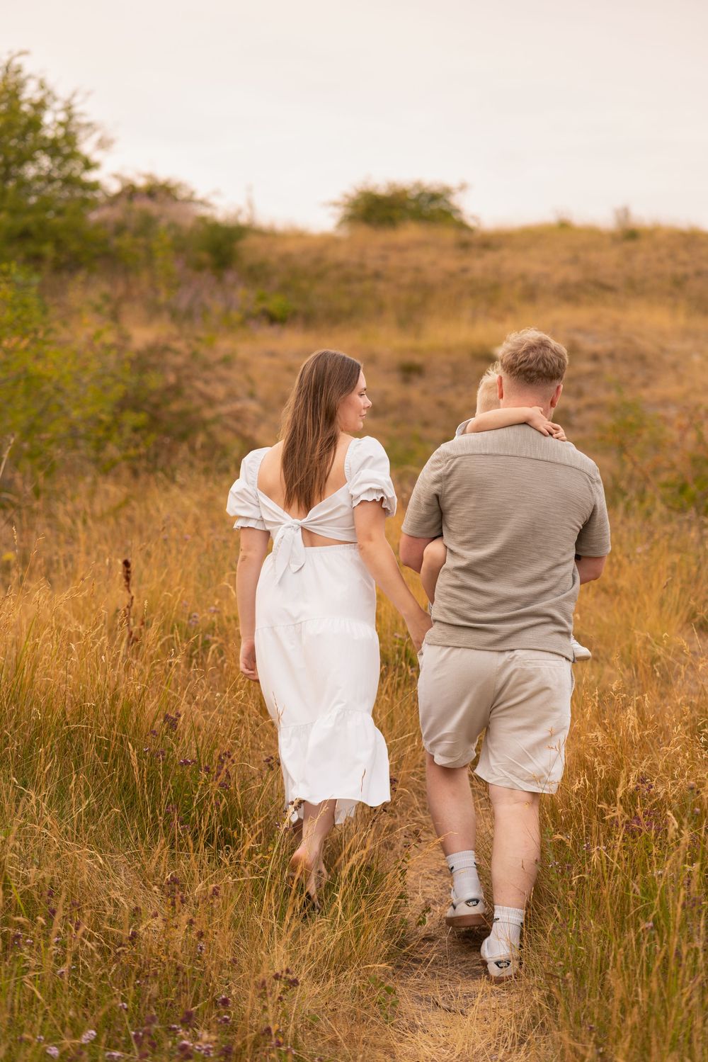 Couple walking hand in hand through golden field, woman in white dress glancing back, man in casual shorts and gray shirt.