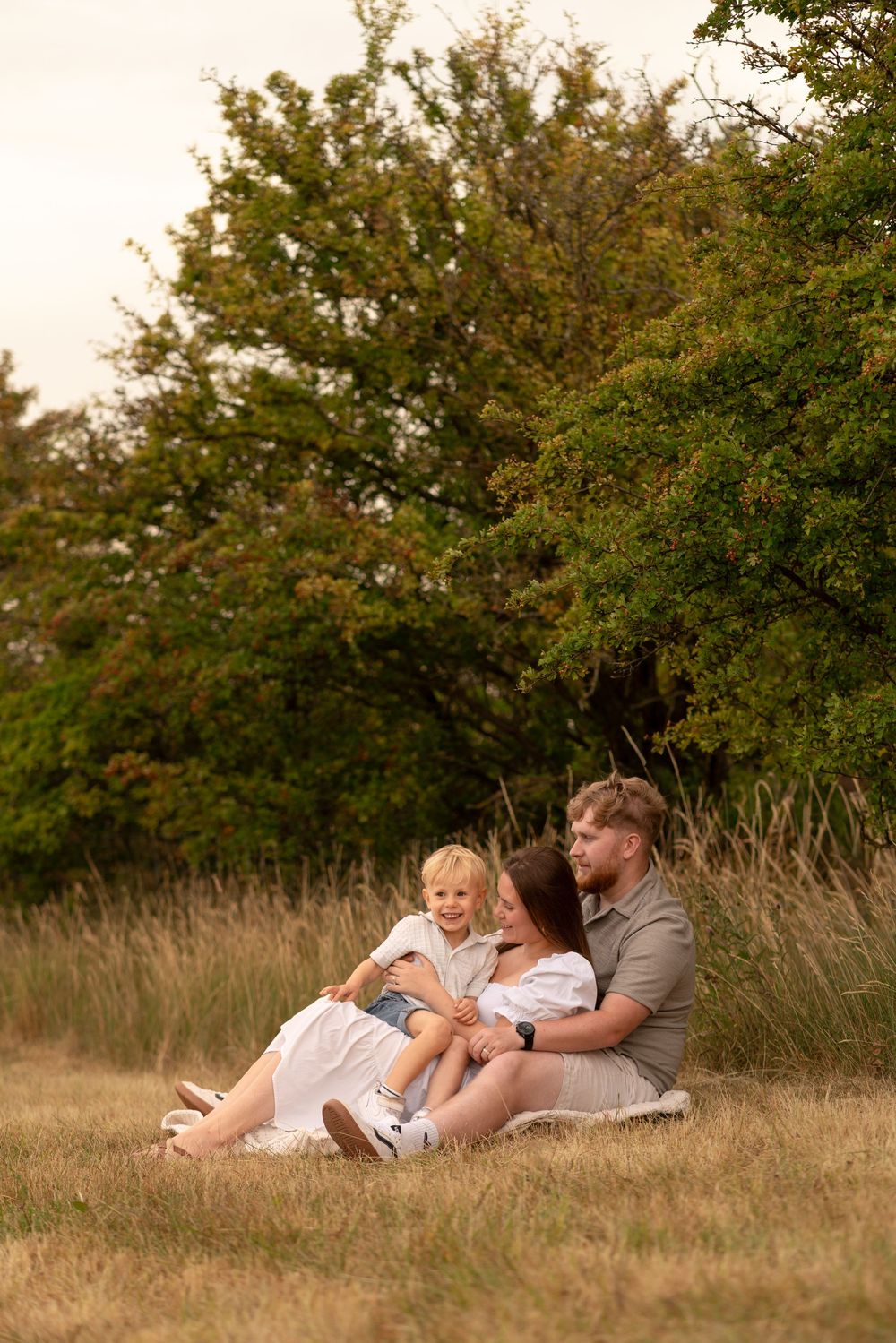 Family of three sitting together on grass field near green trees, parents embracing young blonde child outdoors.