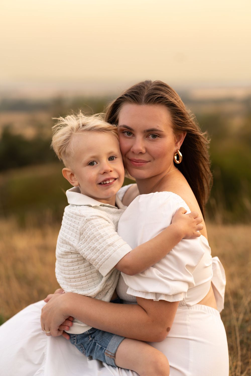 A young mother in a white dress hugs her smiling blonde toddler son in a sunny field at golden hour.