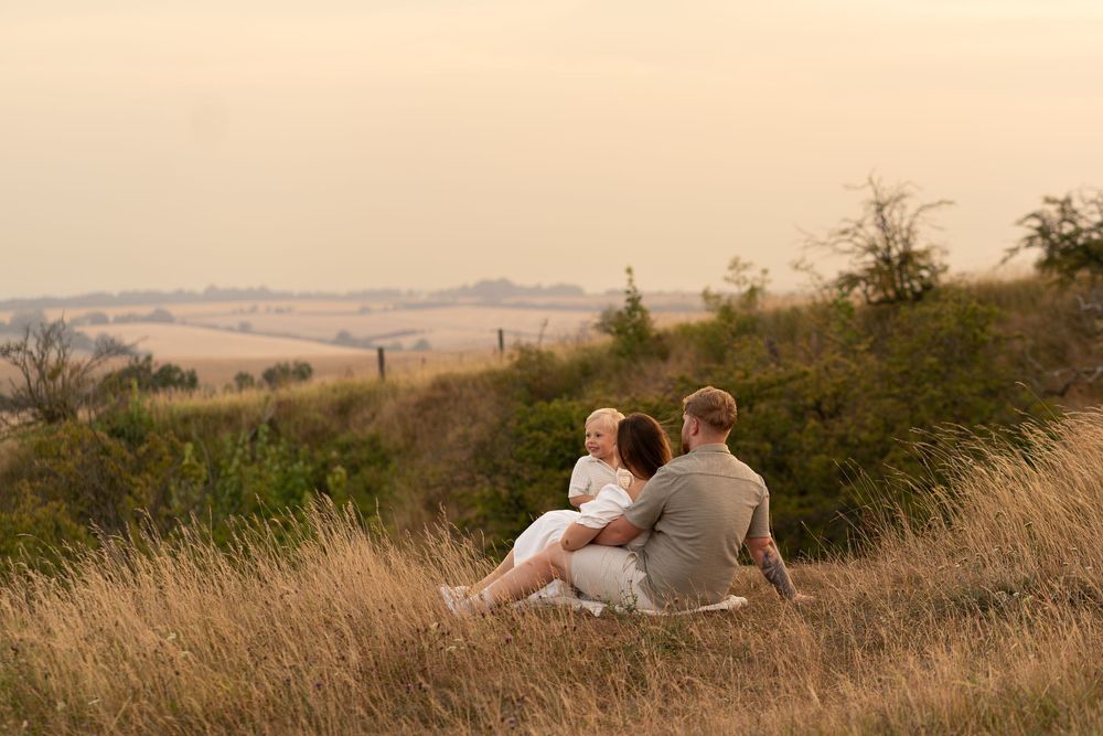 Family sitting together on a grassy hill overlooking golden fields at sunset in a serene countryside landscape.